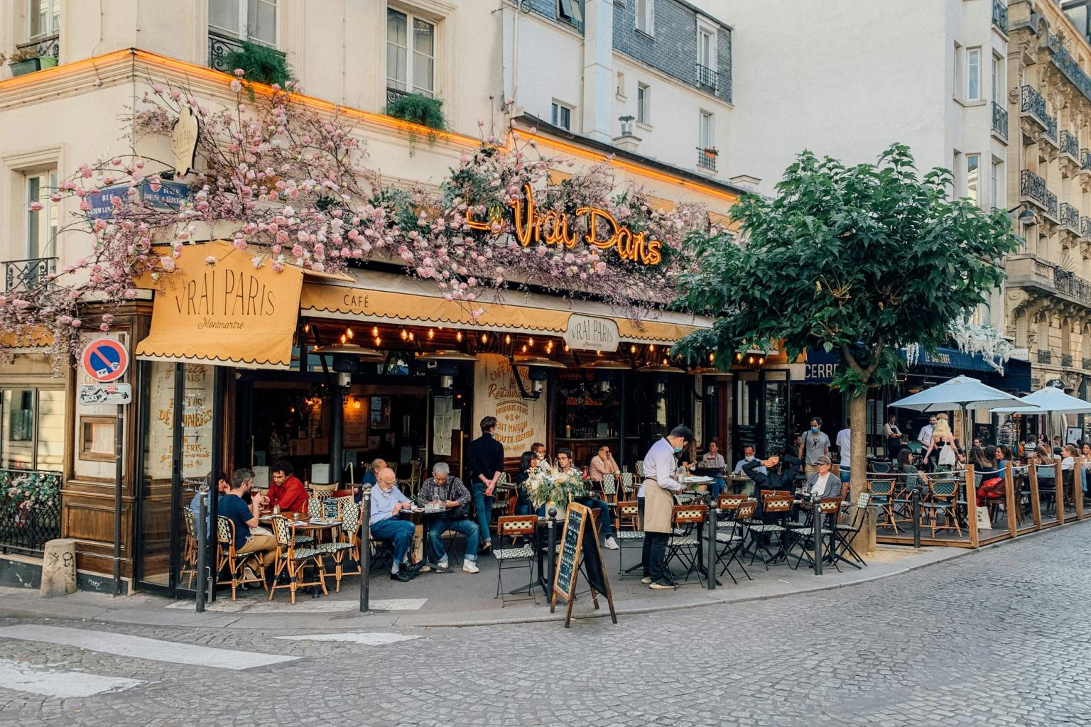 Livlig parisisk uteservering, "Le Vrai Paris", prydd med rosa blommor, med många människor som äter vid utomhusbord på en kullerstensgata.