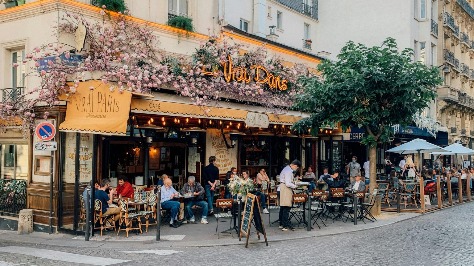 Livlig parisisk uteservering, "Le Vrai Paris", prydd med rosa blommor, med många människor som äter vid utomhusbord på en kullerstensgata.