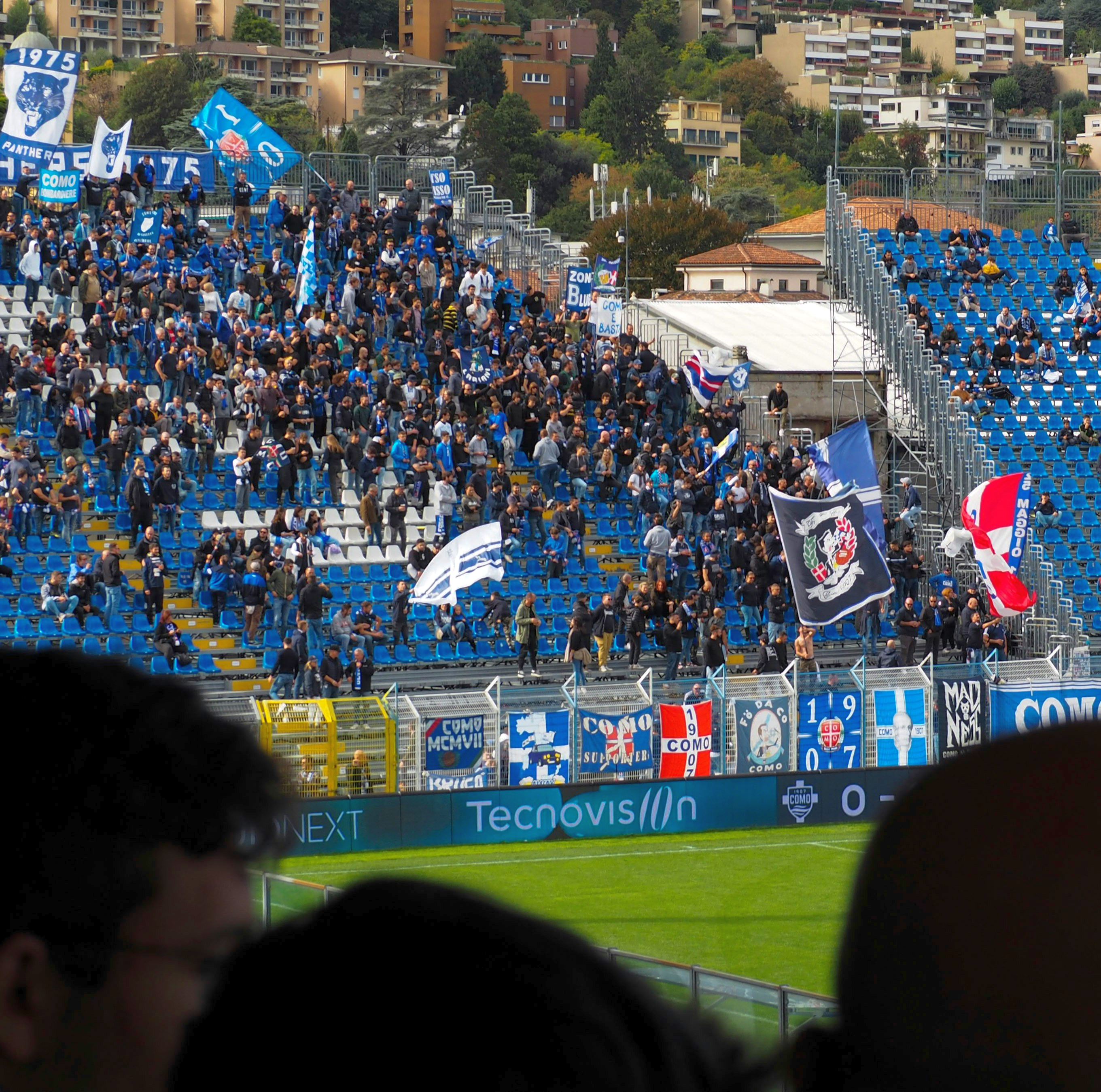 Soccer fans in a stadium stand, many wearing blue, waving flags and banners.
