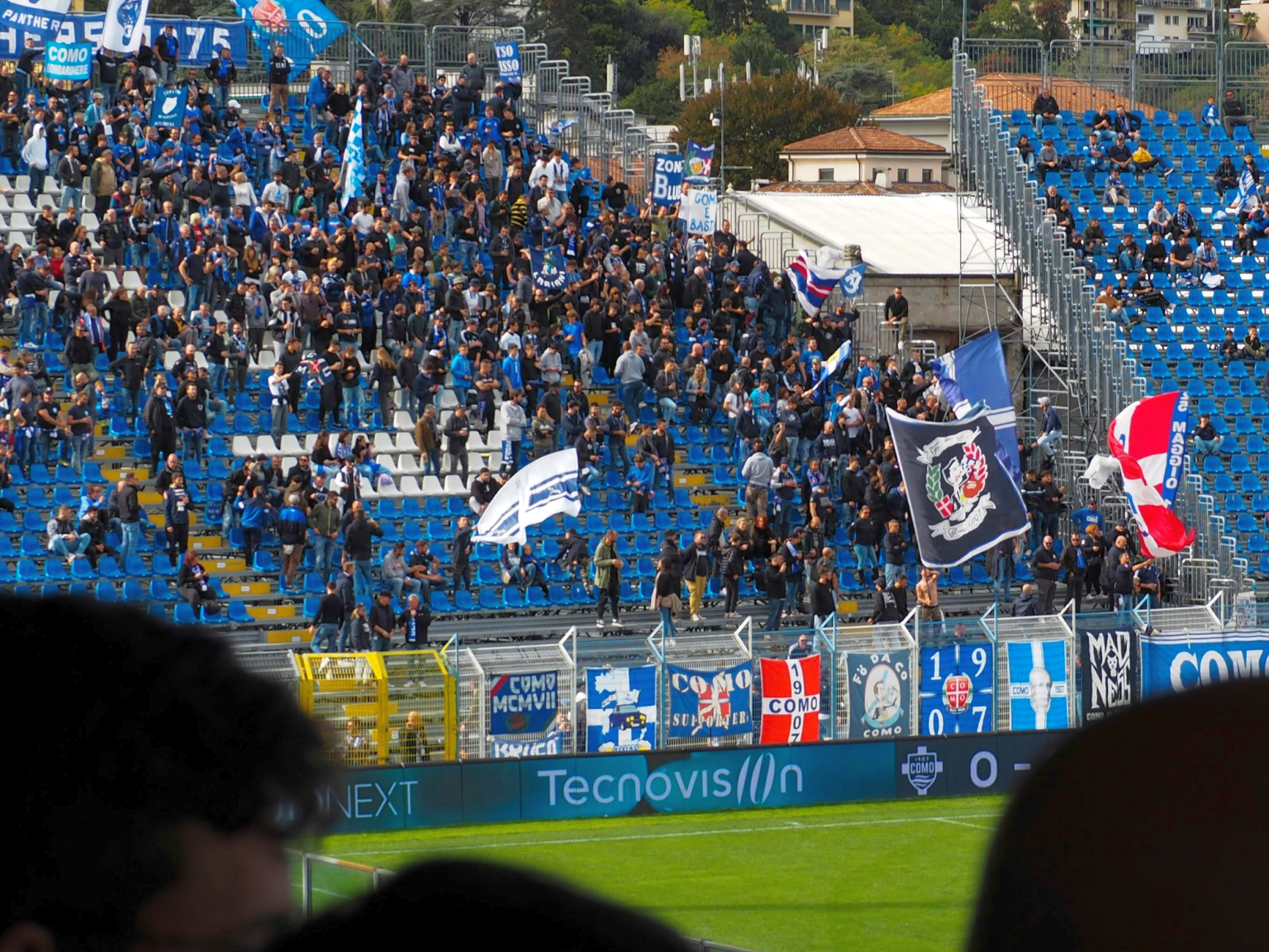 Soccer fans in a stadium stand, many wearing blue, waving flags and banners.