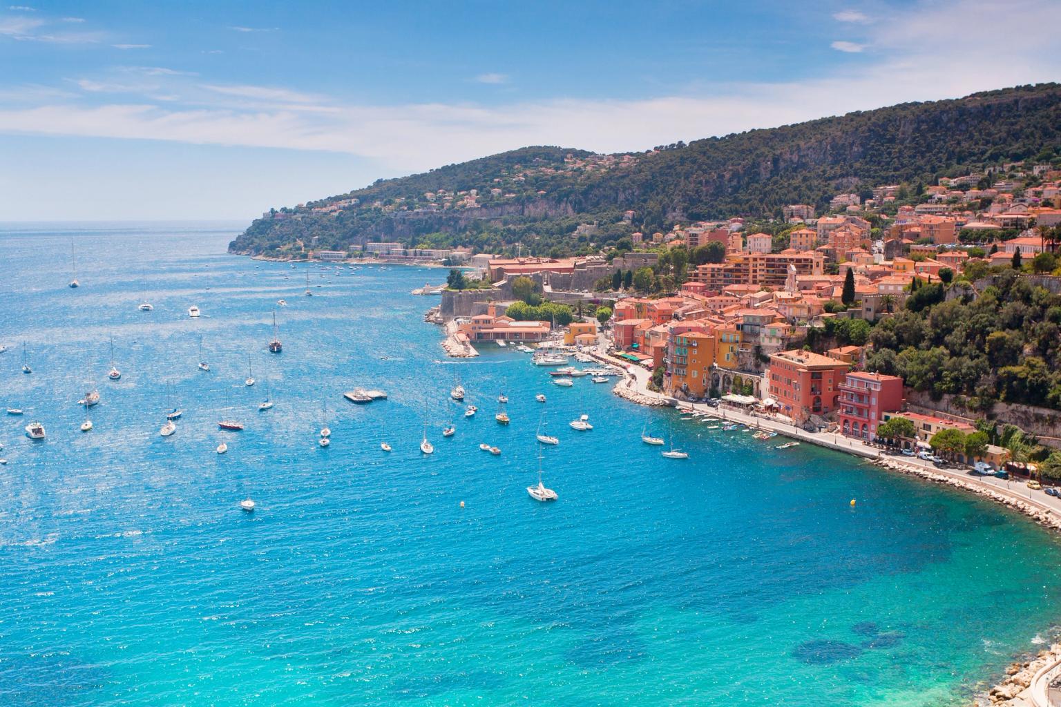 A vibrant coastal town with colorful buildings on a hillside overlooking a clear turquoise bay filled with boats. French Riviera.