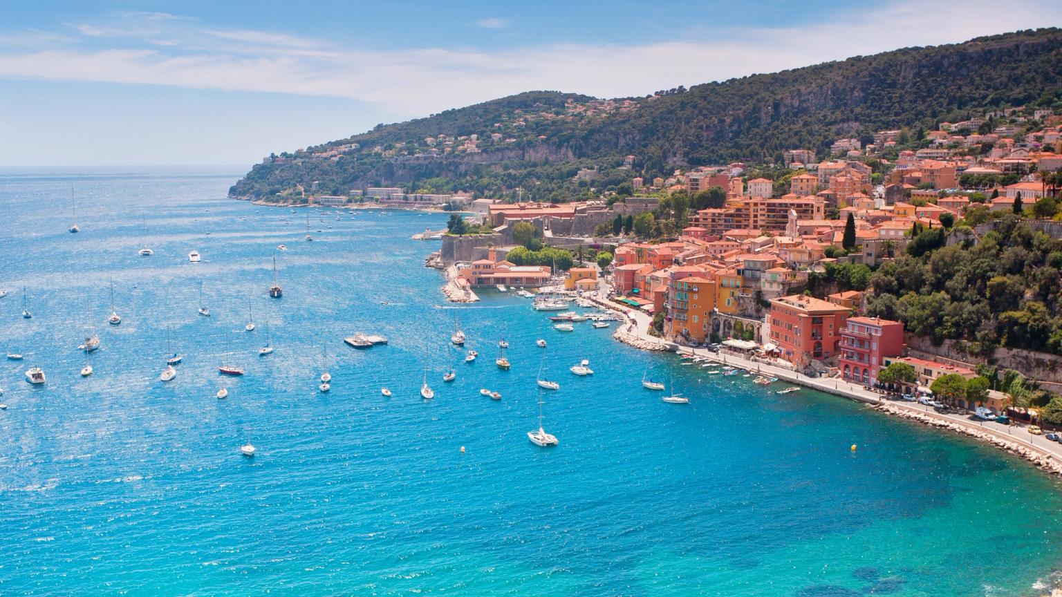 A vibrant coastal town with colorful buildings on a hillside overlooking a clear turquoise bay filled with boats. French Riviera.