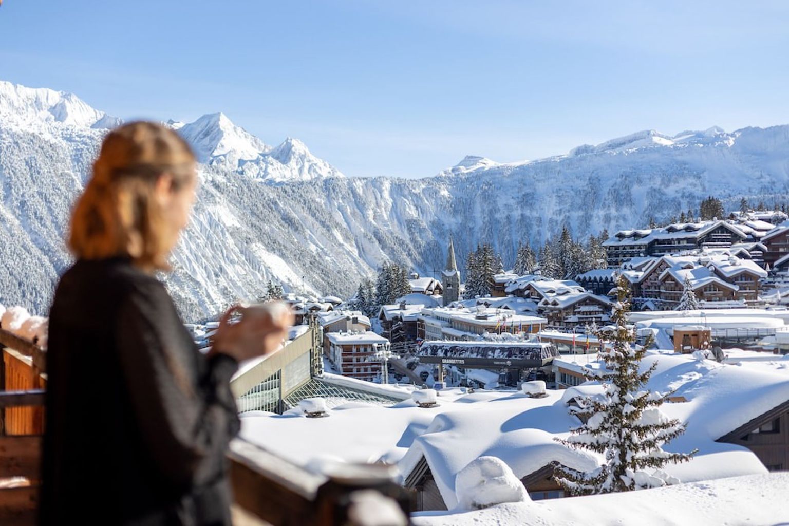 een vrouw drinkt koffie op een balkon met uitzicht op een besneeuwd bergdorp.