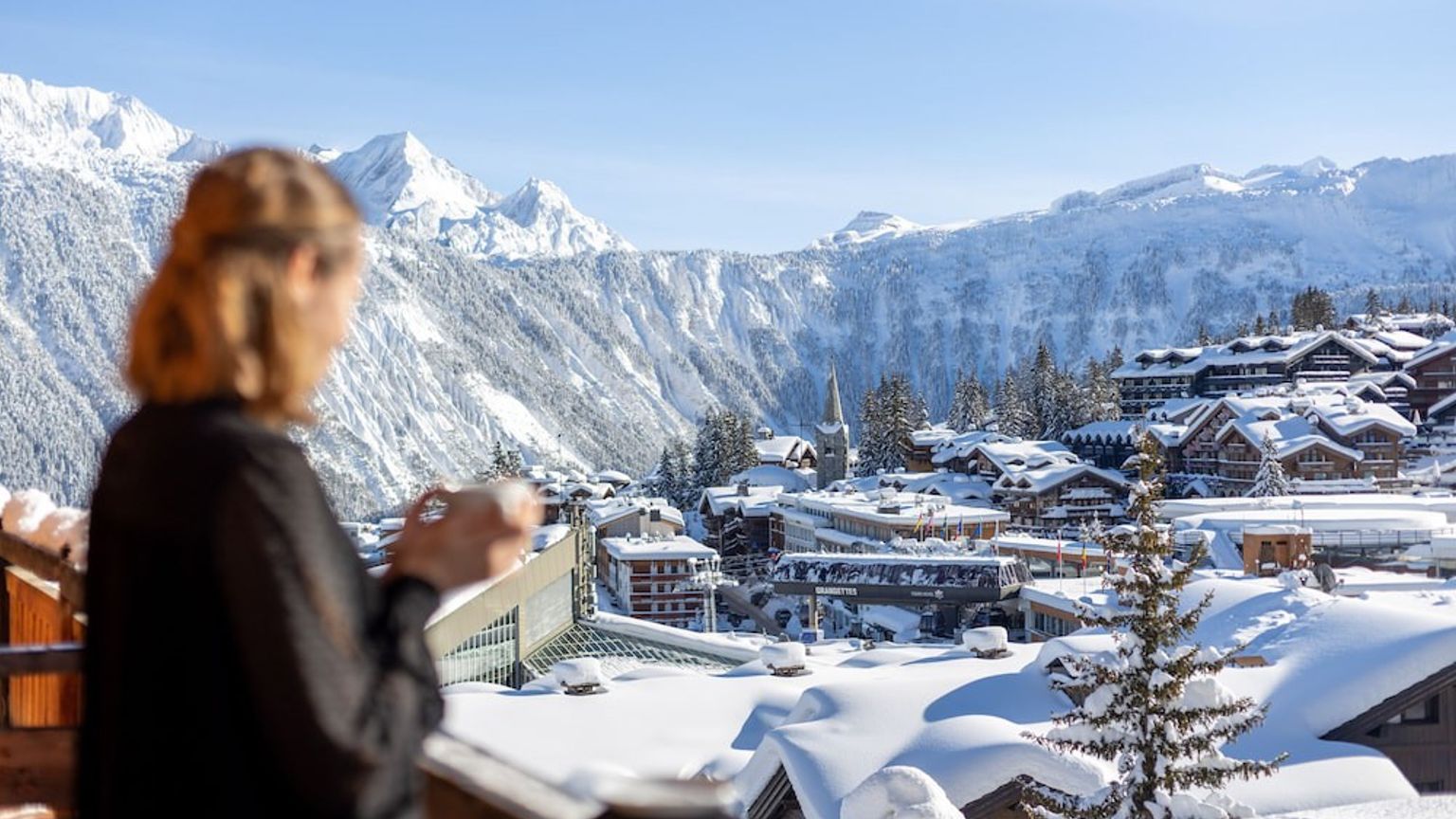 een vrouw drinkt koffie op een balkon met uitzicht op een besneeuwd bergdorp.