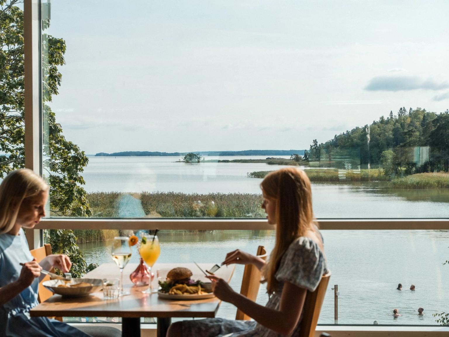 Kaksi naista illastaa suuren ikkunan ääressä, josta on näkymä viehättävälle järvelle, jonka rannat ovat metsän peitossa ja jossa ihmiset uivat.