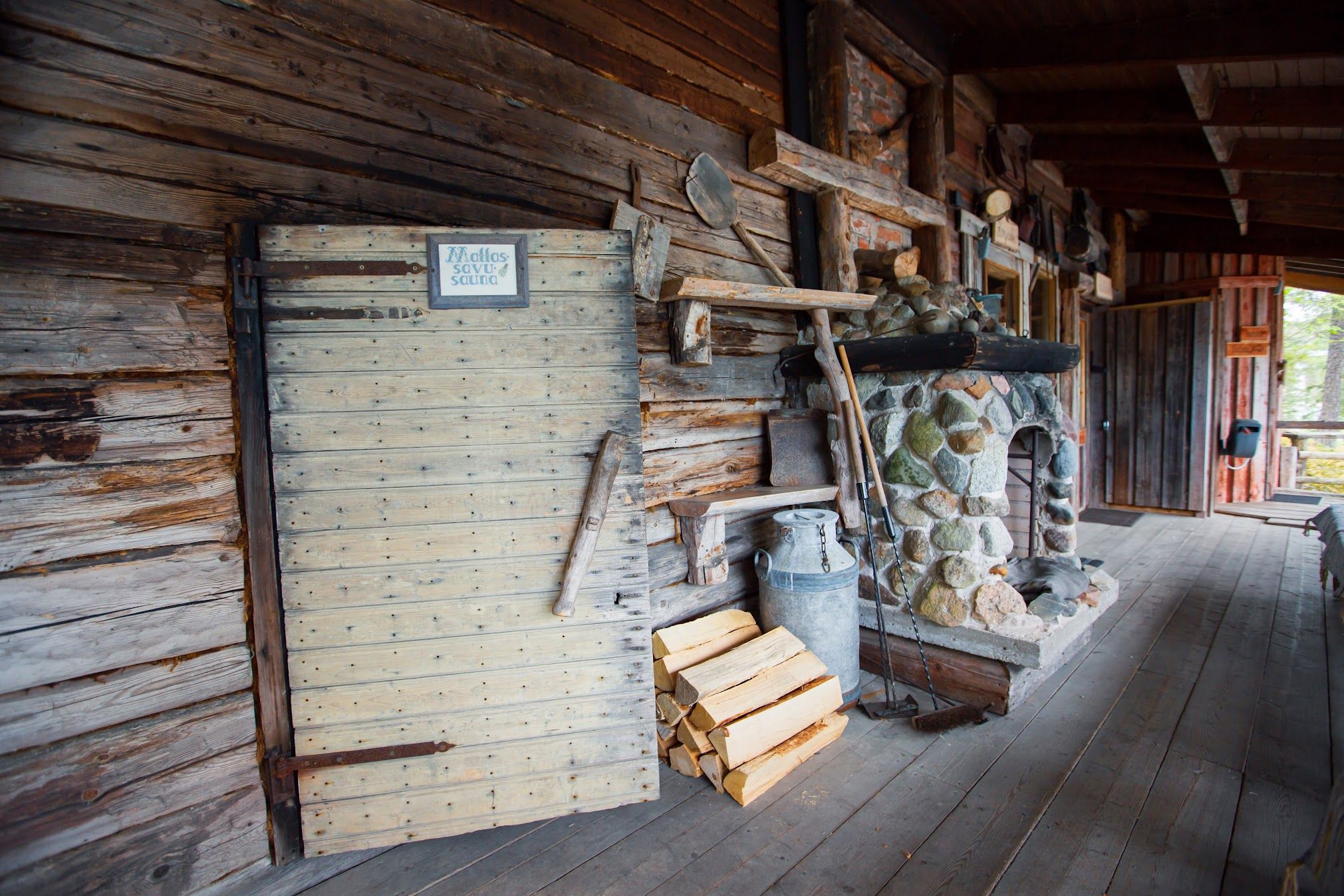 Rustik træhyttes veranda med en stenpejs, stablet brænde og en stor plankedør.