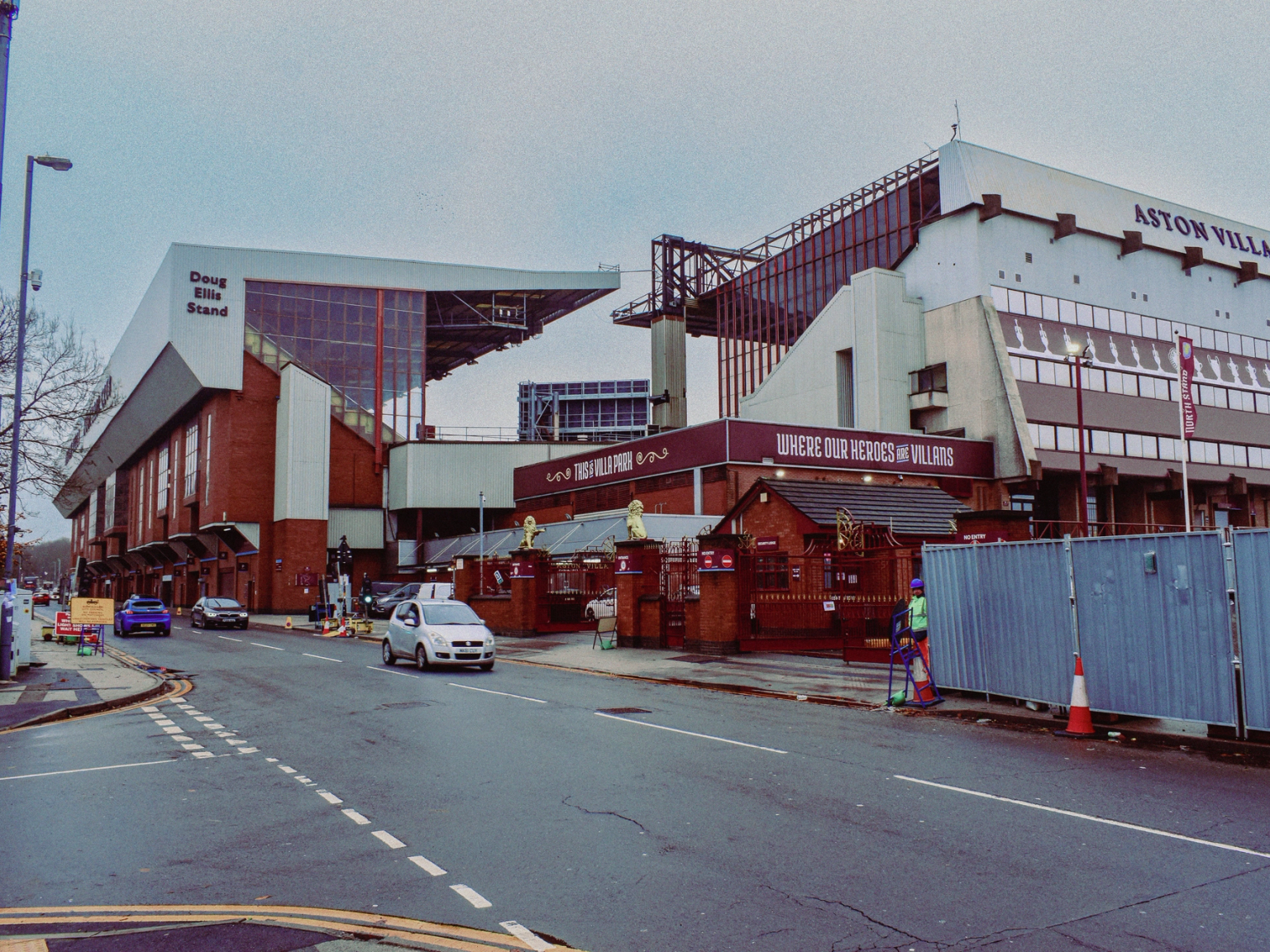 Exterior view of Aston Villa Football Club stadium, including the Doug Ellis Stand, on an overcast day with a street and cars in the foreground.