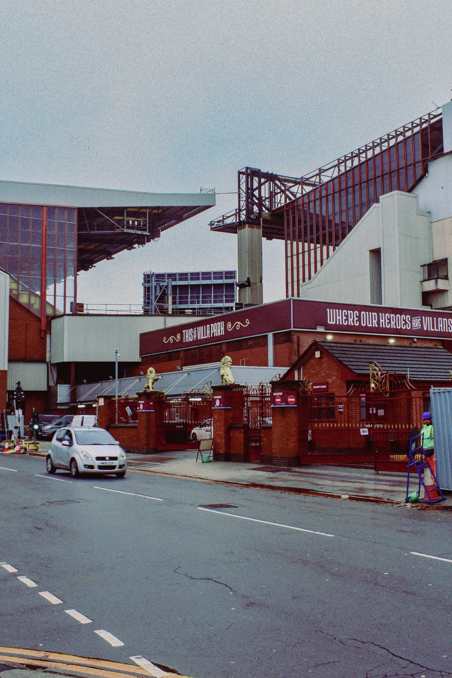 Exterior view of Aston Villa Football Club stadium, including the Doug Ellis Stand, on an overcast day with a street and cars in the foreground.