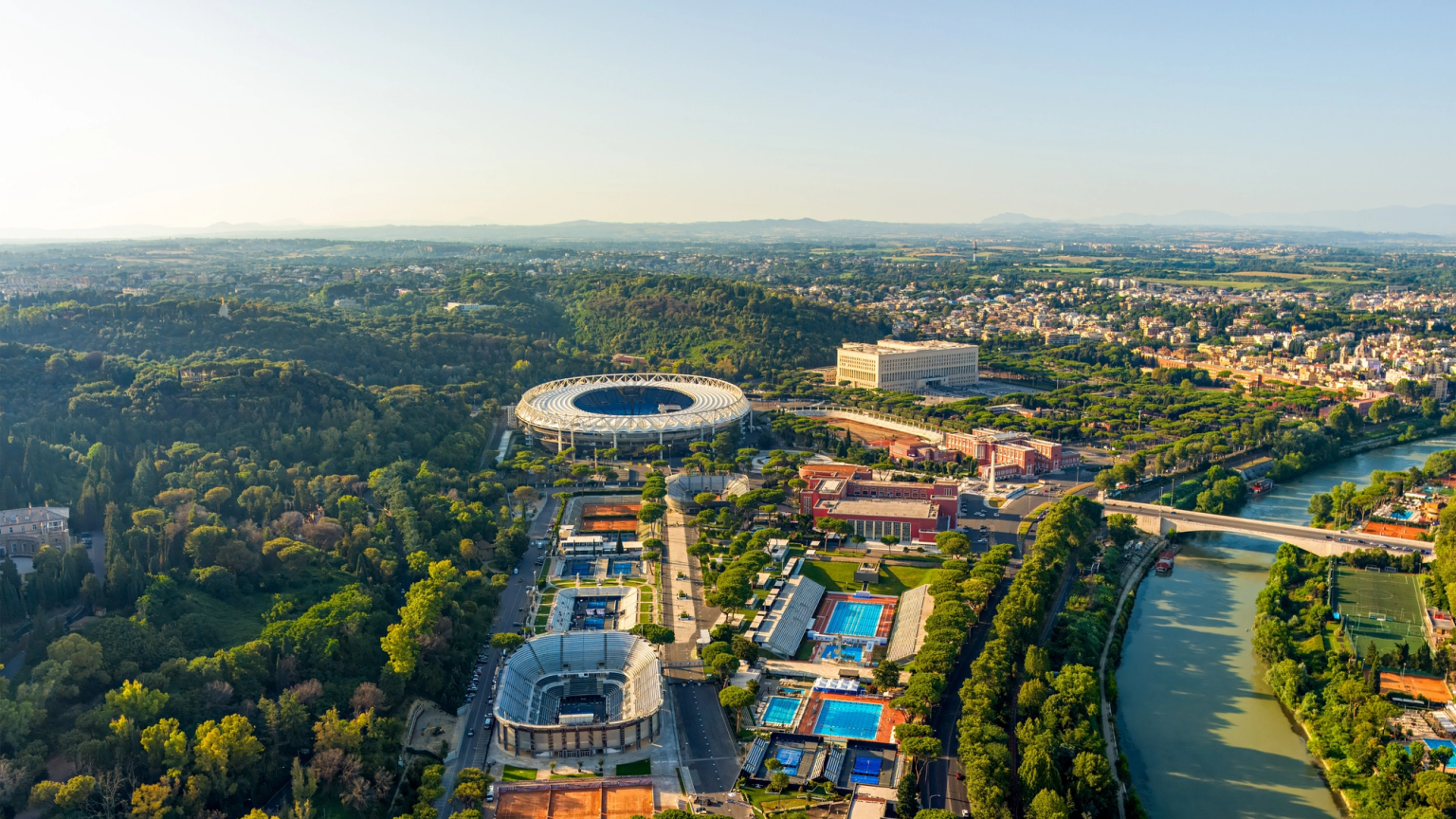 Aerial view of Stadio Olimpico, a large sports complex, and the Tiber River in a green urban landscape.