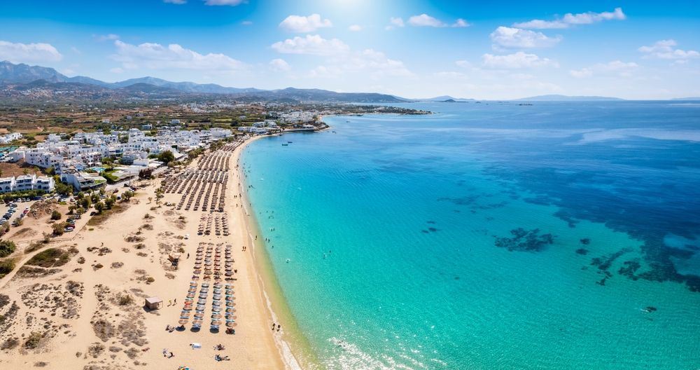 Aerial view of a sunny beach with rows of lounge chairs, white buildings, and clear turquoise ocean. Naxos, Greece