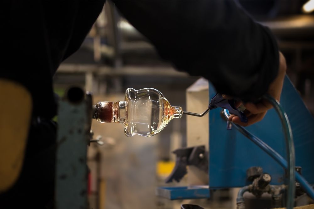 En hånd bruker et munnstykkeverktøy for å forme et klart glassobjekt på en roterende spindel.