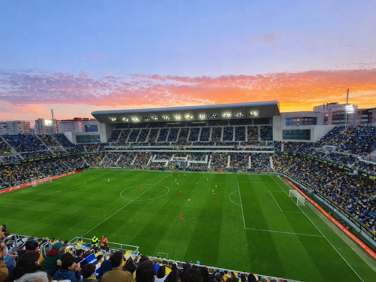 Um céu vibrante de pôr do sol sobre um estádio de futebol lotado durante uma partida.