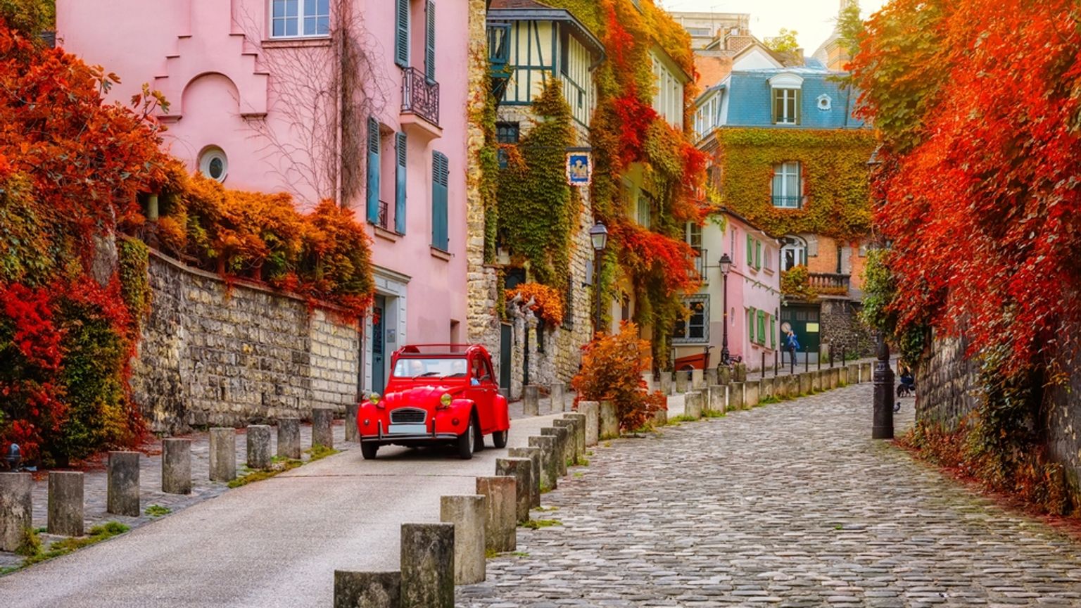 Een schilderachtige straat omzoomd met gebouwen bedekt met levendig rood en oranje herfstgebladerte, met een rode vintage auto. Parijs
