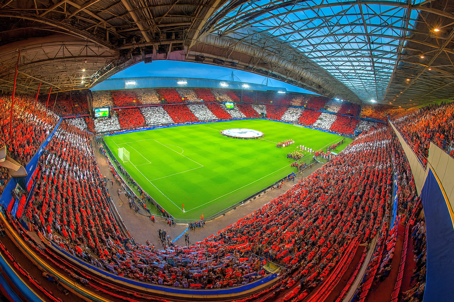 Eine Draufsicht auf ein Fußballstadion, das mit Fans gefüllt ist, die rote und weiße Muster auf den Tribünen um ein grünes Spielfeld herum bilden.