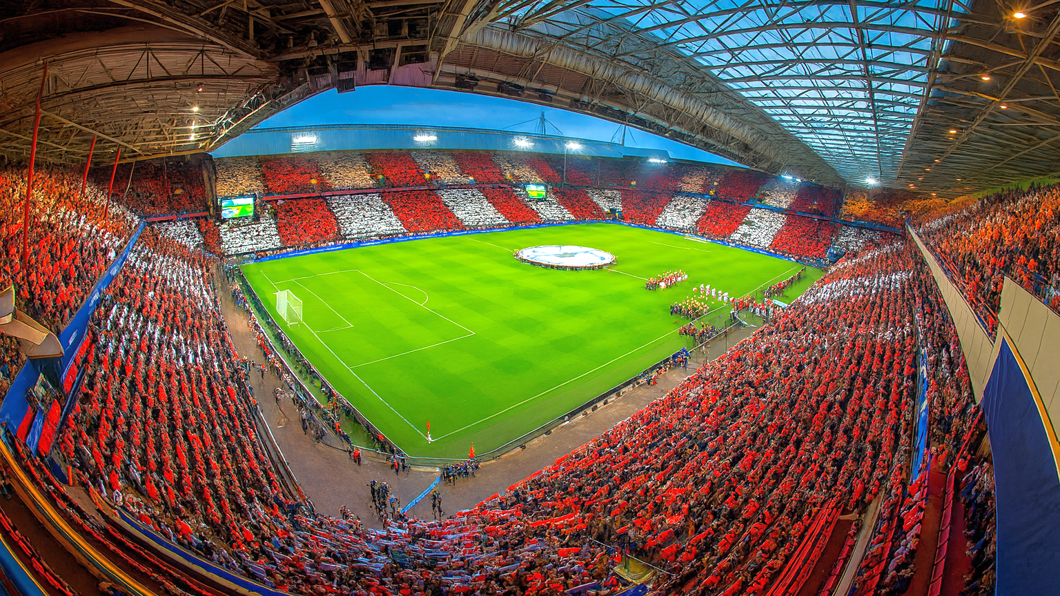 Eine Draufsicht auf ein Fußballstadion, das mit Fans gefüllt ist, die rote und weiße Muster auf den Tribünen um ein grünes Spielfeld herum bilden.
