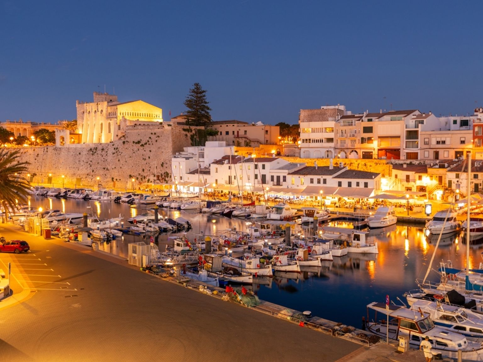 En livlig hamn i skymningen med upplysta strandbyggnader, många båtar och en gammal fästning. Ciutadella, Menorca