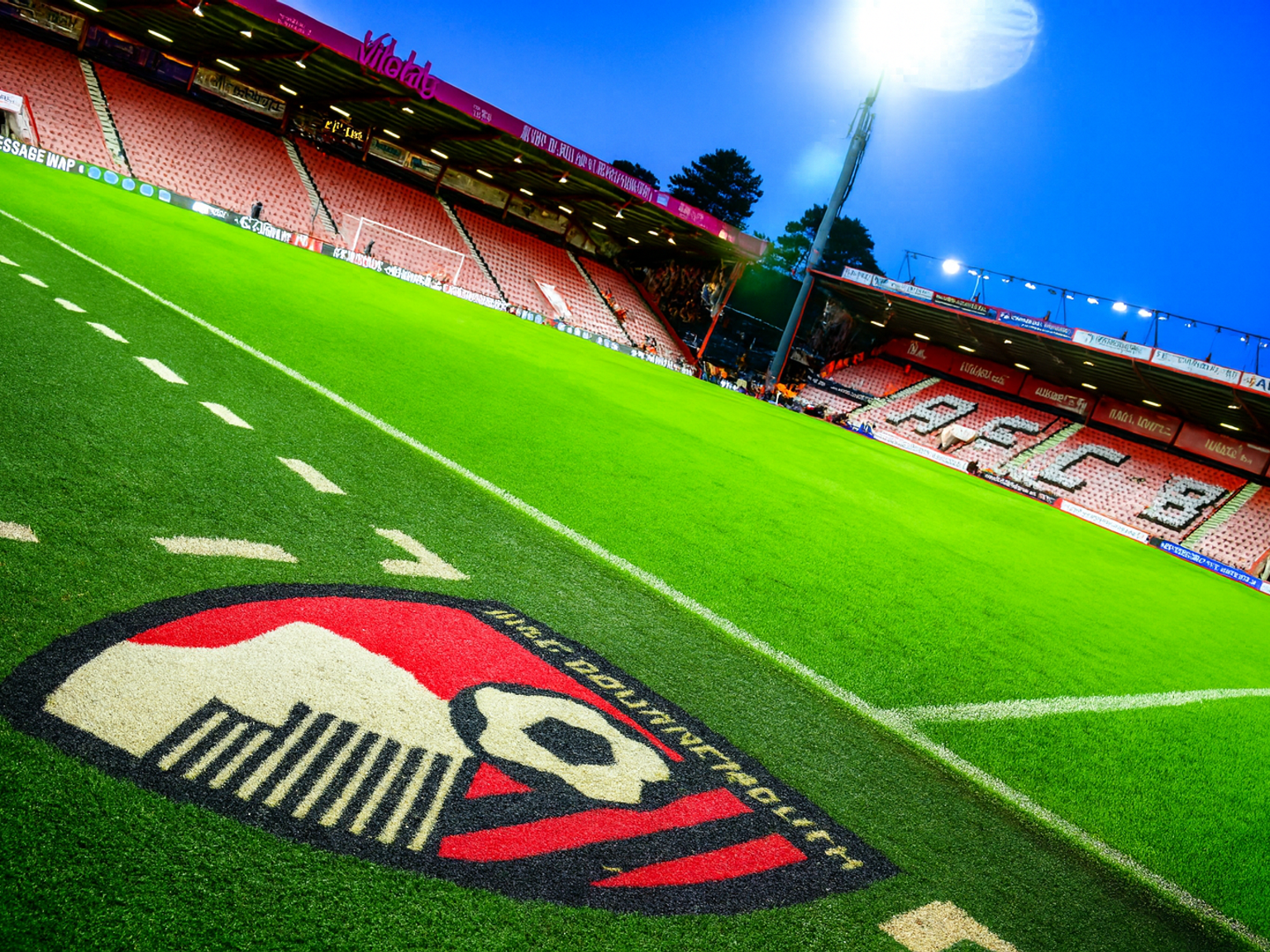 Fotballbane med AFC Bournemouth-logo, røde stadionseter og sterkt flomlys.