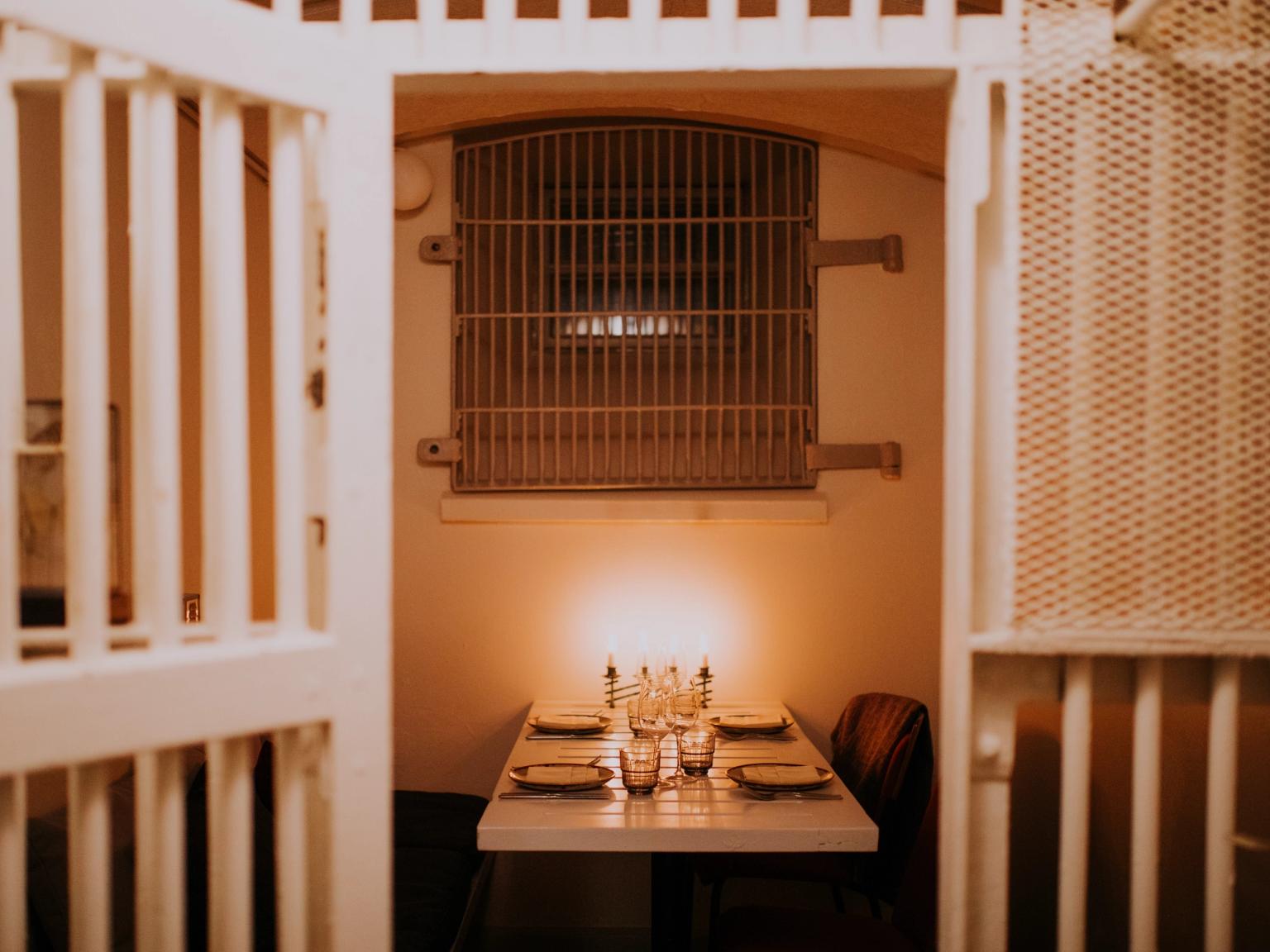 Kynttilänvalossa katettu ruokapöytä neljälle sellimäisessä huoneessa, jossa on kalteri-ikkuna.