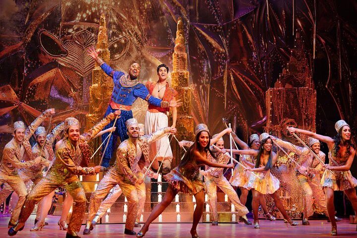 Un spectacle de scène vibrant avec le Génie en bleu et Aladdin en blanc, entourés de nombreux danseurs en costumes scintillants sur un grand décor doré.
