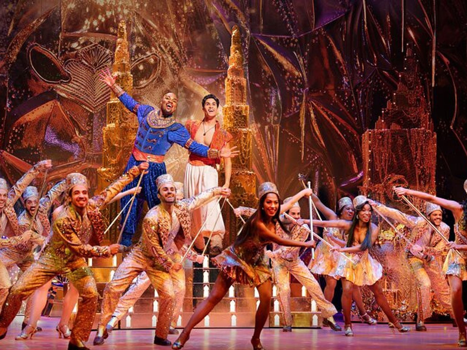 Un spectacle de scène vibrant avec le Génie en bleu et Aladdin en blanc, entourés de nombreux danseurs en costumes scintillants sur un grand décor doré.