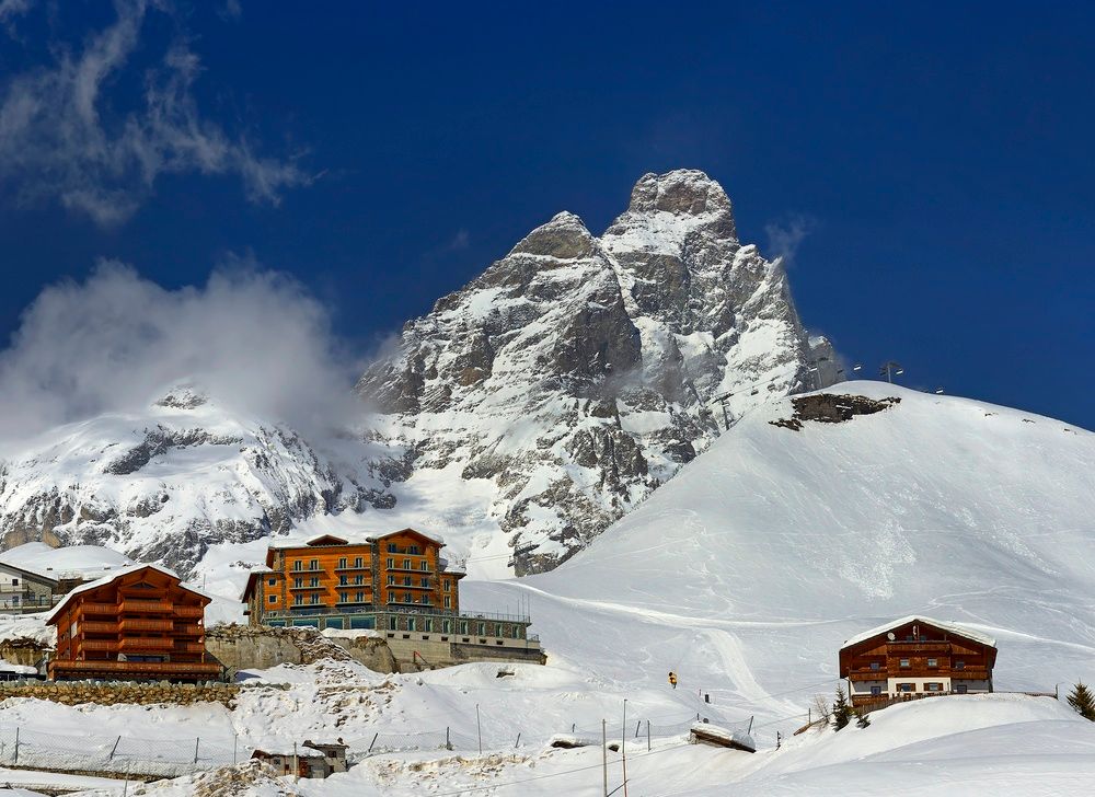 En fremtredende snødekt fjelltopp ruver over et skianlegg med et oransje hotell og hytter under en dypblå himmel.