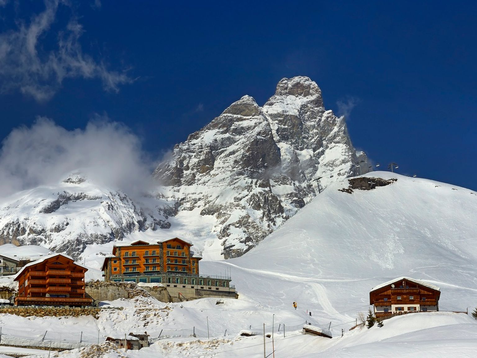 En fremtredende snødekt fjelltopp ruver over et skianlegg med et oransje hotell og hytter under en dypblå himmel.