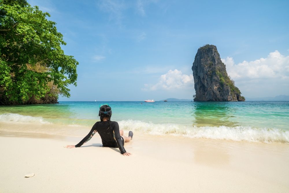 Person i rashguard och snorkelmask som sitter på en vit sandstrand, vänd mot ett turkost hav med en stor klippö i fjärran.