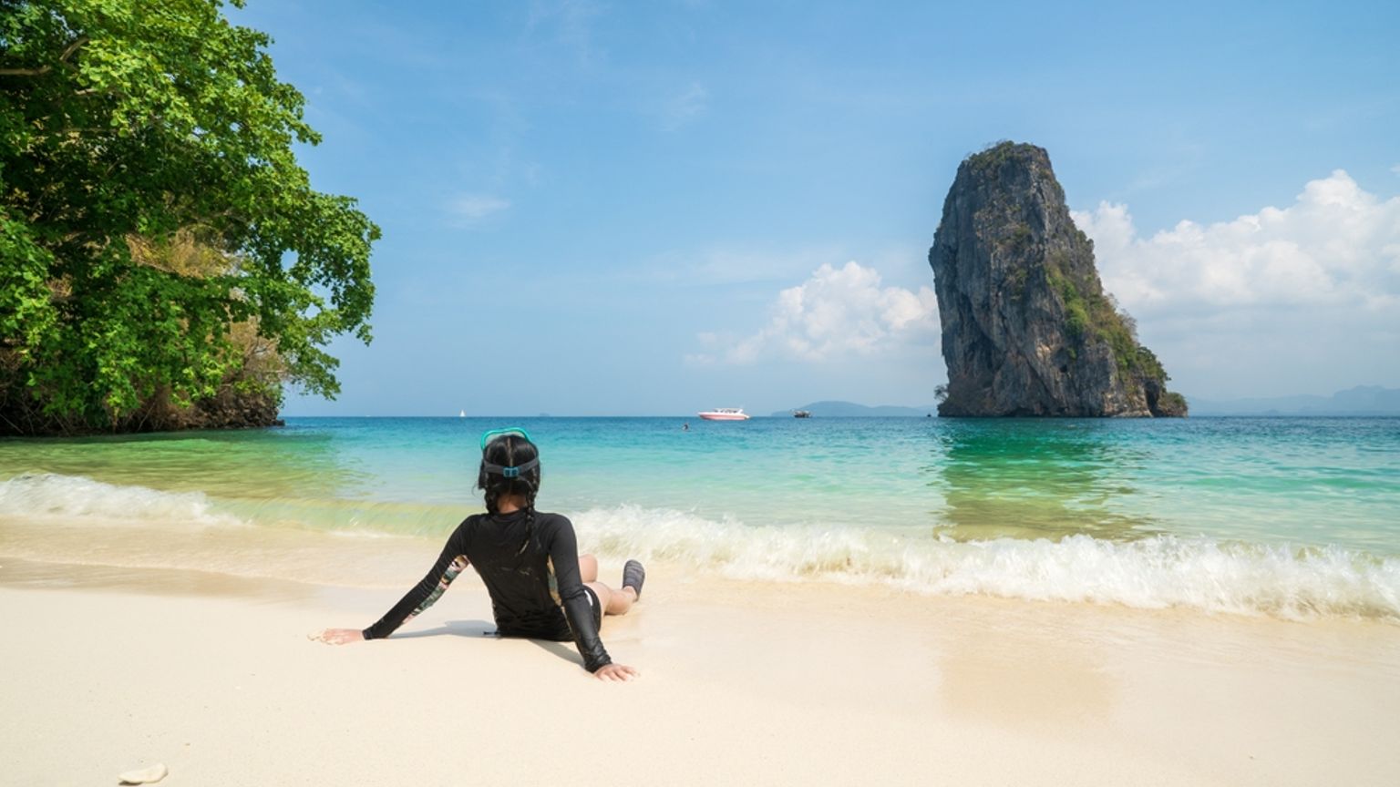 Person i rashguard och snorkelmask som sitter på en vit sandstrand, vänd mot ett turkost hav med en stor klippö i fjärran.