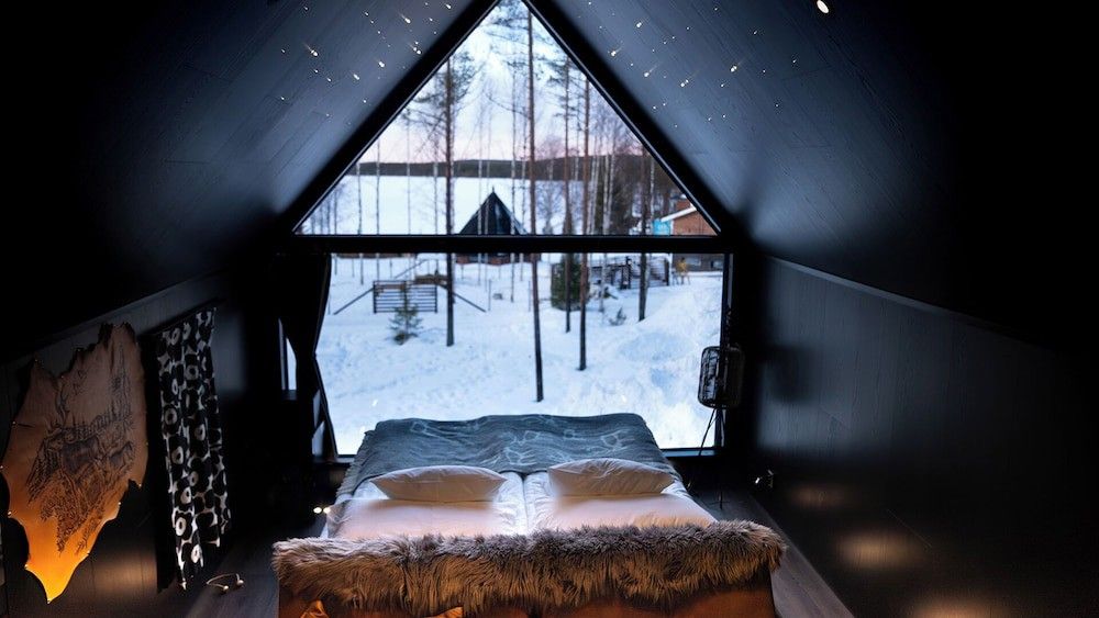 Ett mörkt sovrum med en säng som vetter mot ett stort triangulärt fönster med utsikt över en snöig skog och sjö, under ett tak med stjärnliknande ljus. Aito Igloo & Spa Resort