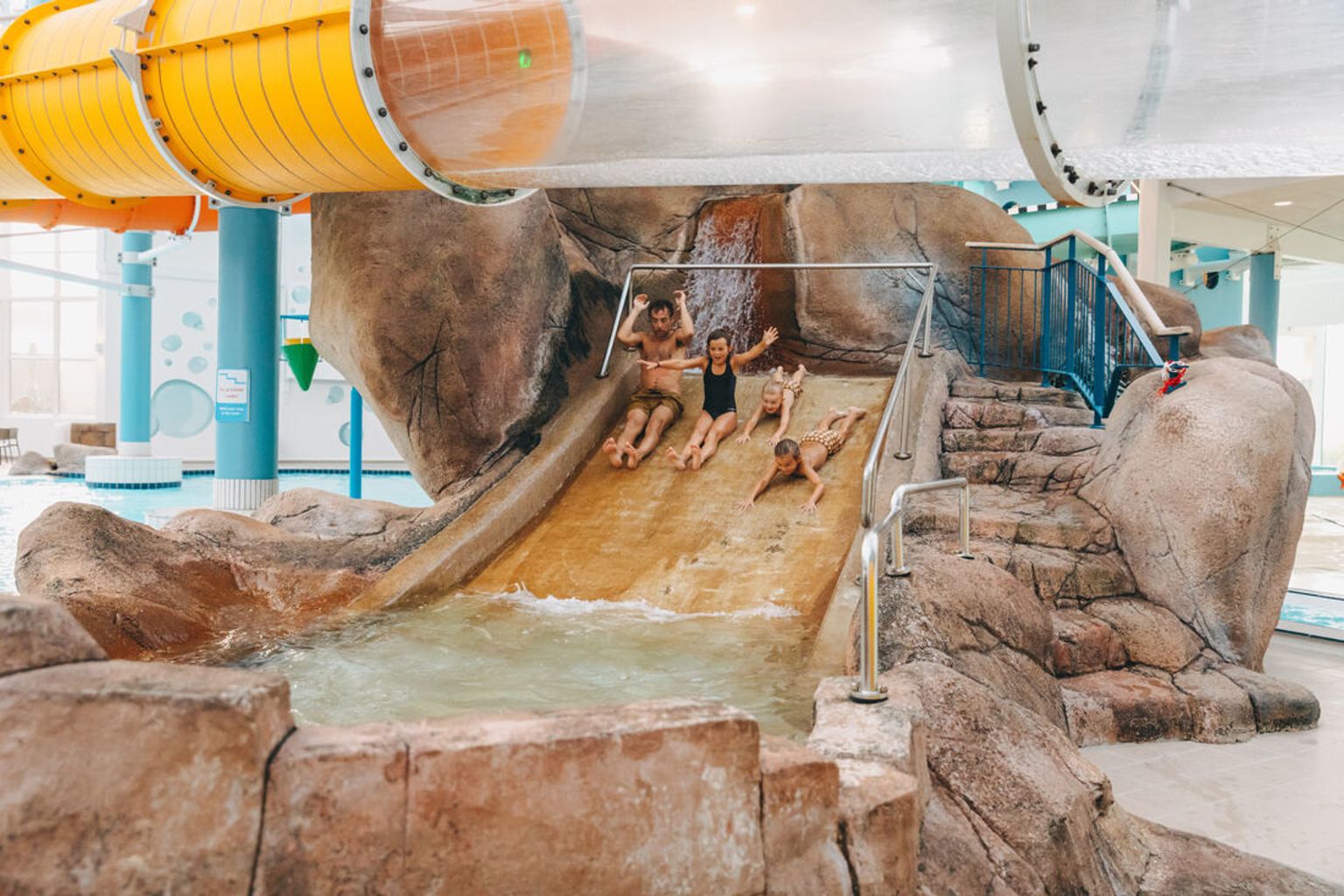 En man och tre barn ler medan de åker nerför en klippformad vattenrutschbana på en inomhusvattenpark.