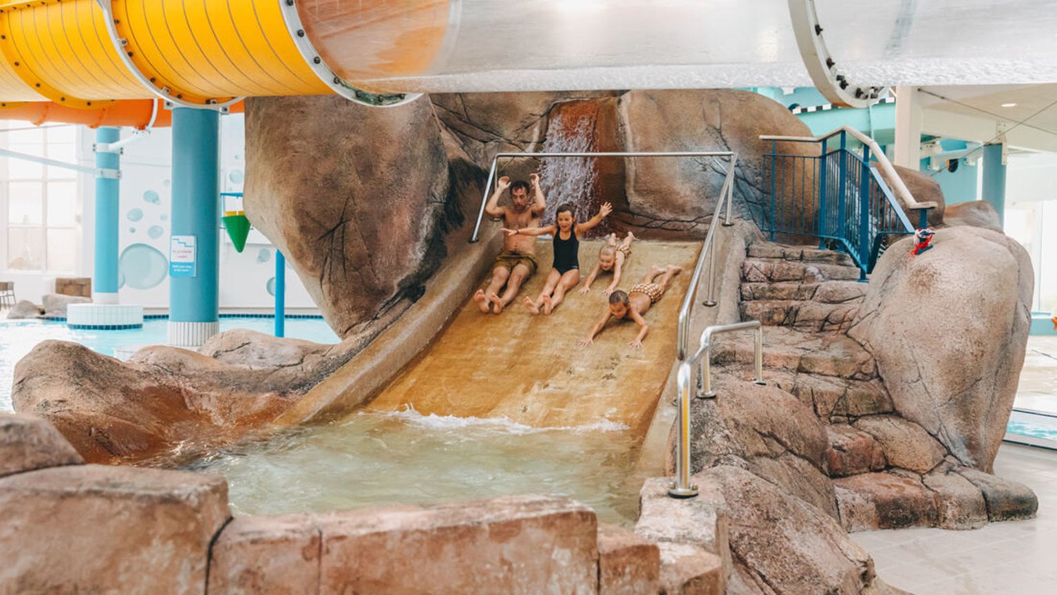 En man och tre barn ler medan de åker nerför en klippformad vattenrutschbana på en inomhusvattenpark.