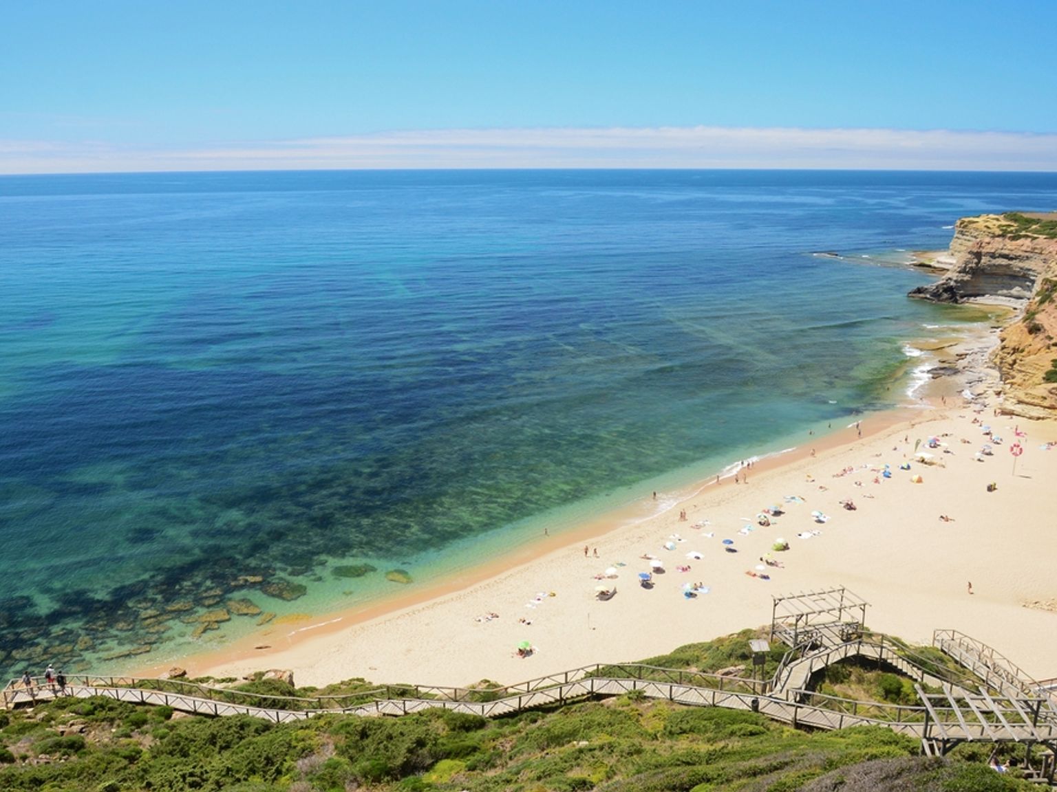 Flygfoto av en solig strand med klart turkost vatten, gyllene sand och en slingrande träpromenad från klippor. Ericeira, Portugal