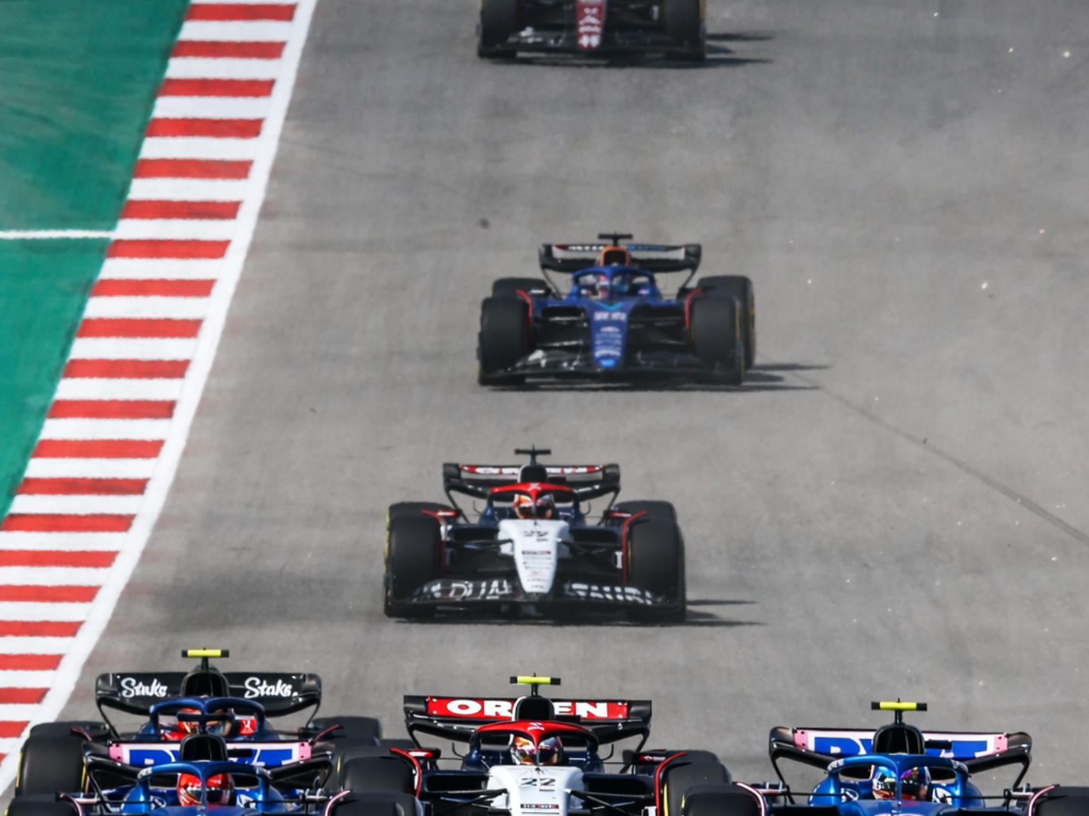 Multiple Formula 1 cars race in a line on a winding track with striped curbs.