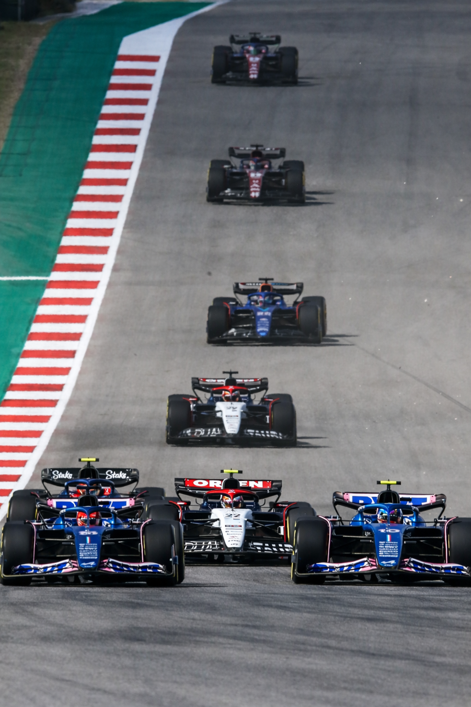 Multiple Formula 1 cars race in a line on a winding track with striped curbs.