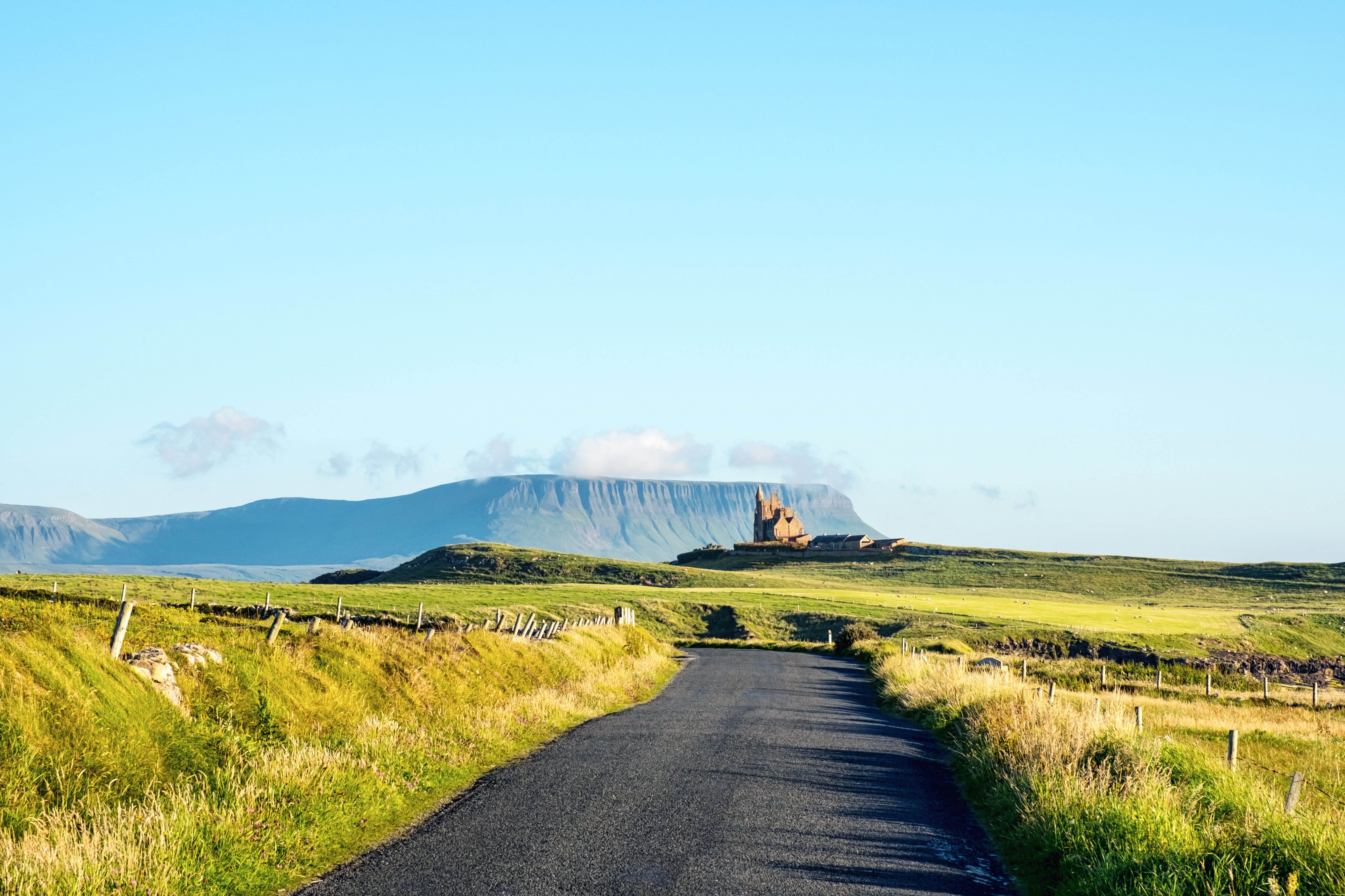 A winding road through green fields towards a distant castle ruin and flat-topped mountain under a blue sky.