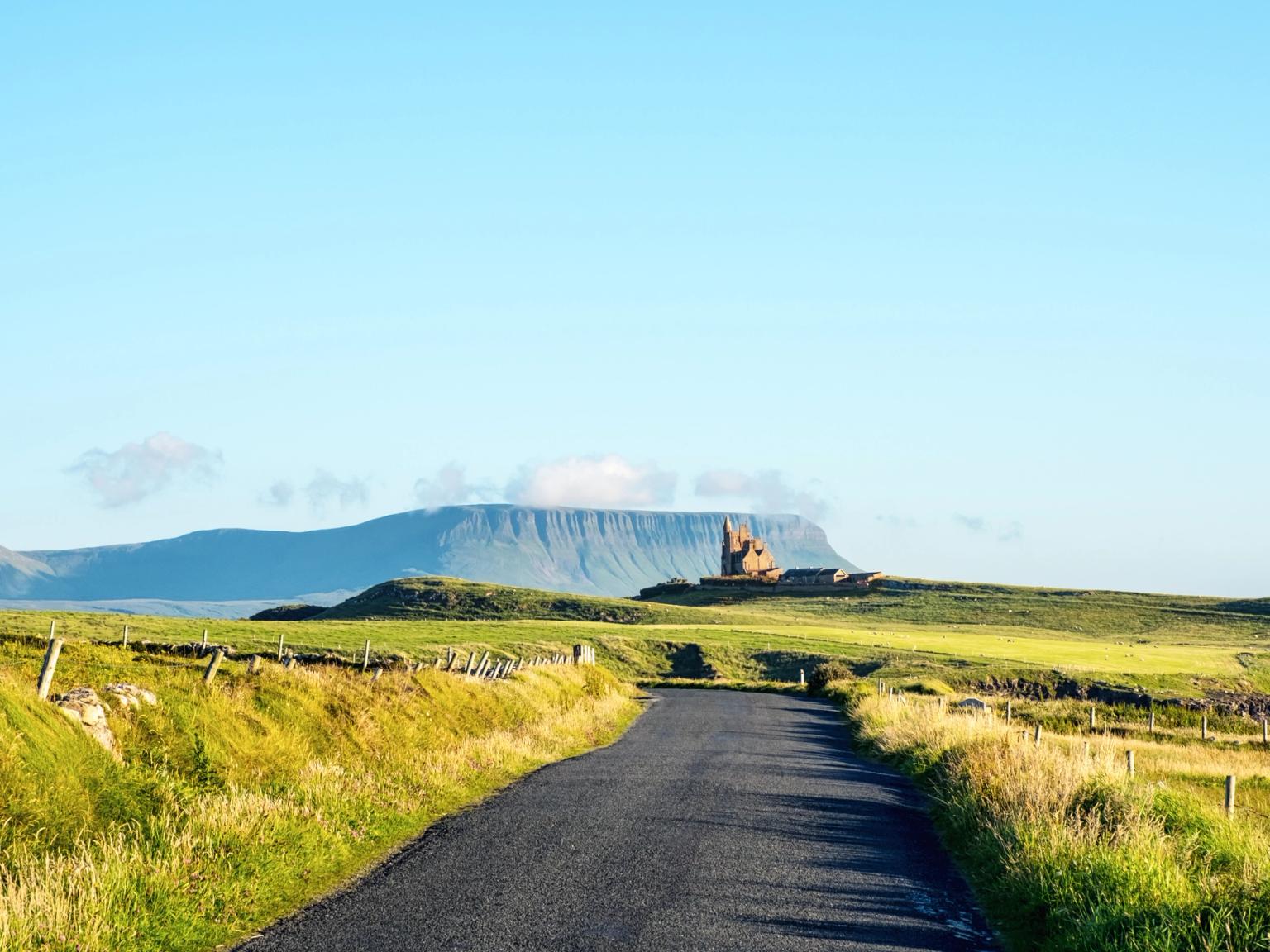 A winding road through green fields towards a distant castle ruin and flat-topped mountain under a blue sky.