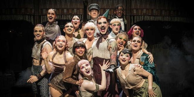 Duży, uśmiechnięty zespół wykonawców w teatralnych kostiumach i z dramatycznym makijażem.