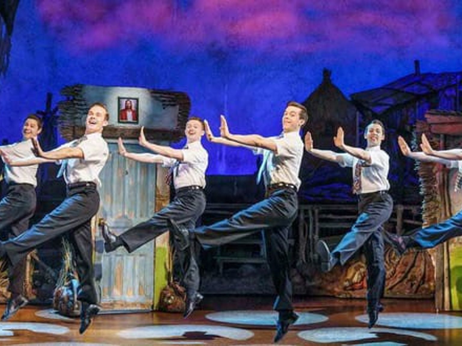 Seks menn i hvite skjorter og slips som danser i takt på en teaterscene.