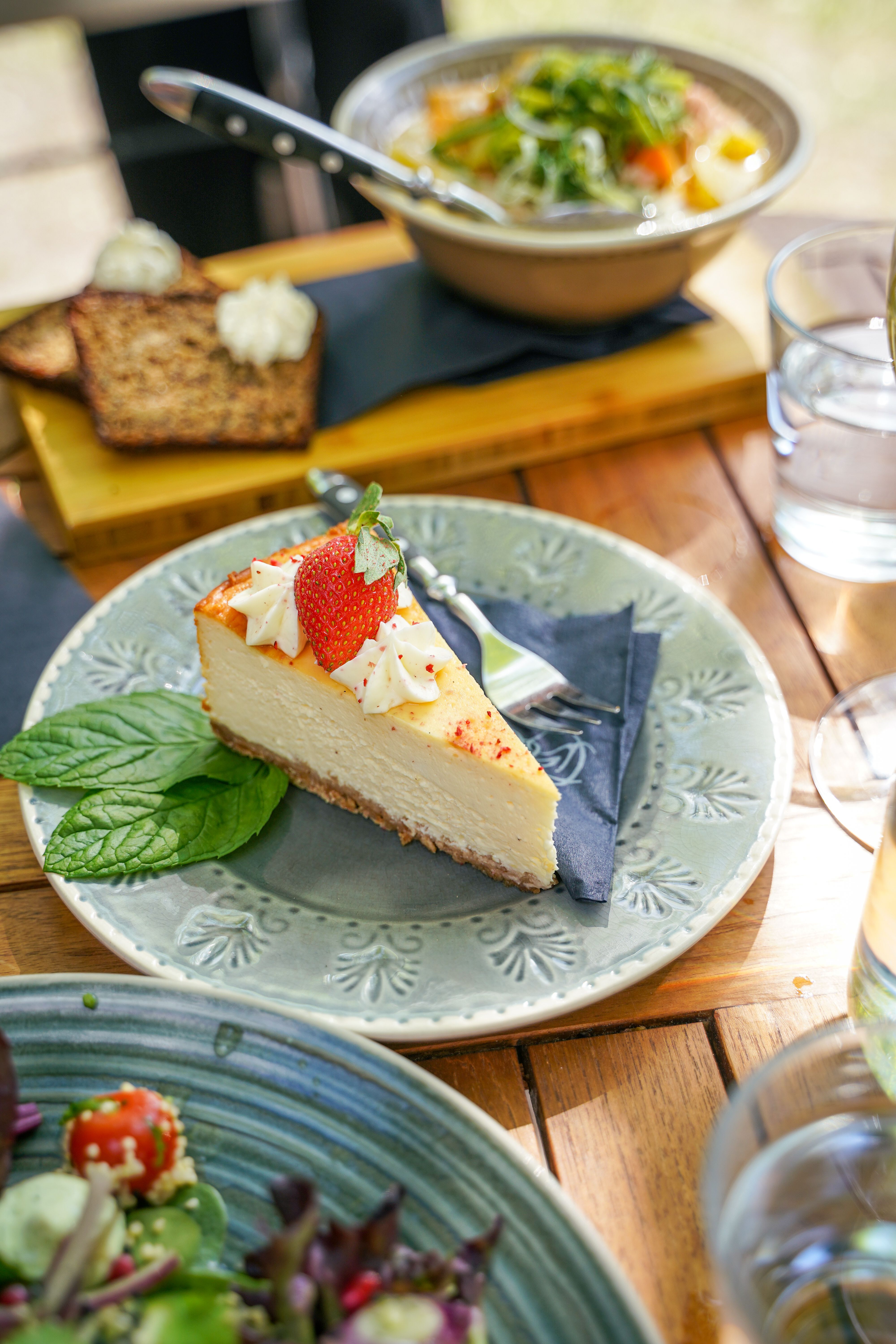 Et stykke cheesecake toppet med et jordbær, flødeskum og røde krymmel, serveret på en mønstret tallerken med mynteblade. Andre retter er slørede i baggrunden på et træbord.