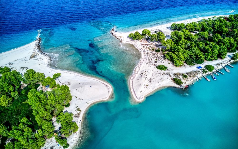 Luftfoto af en smal turkis kanal, der skærer gennem en hvid sandtange, flankeret af grønne træer og en bådebro.