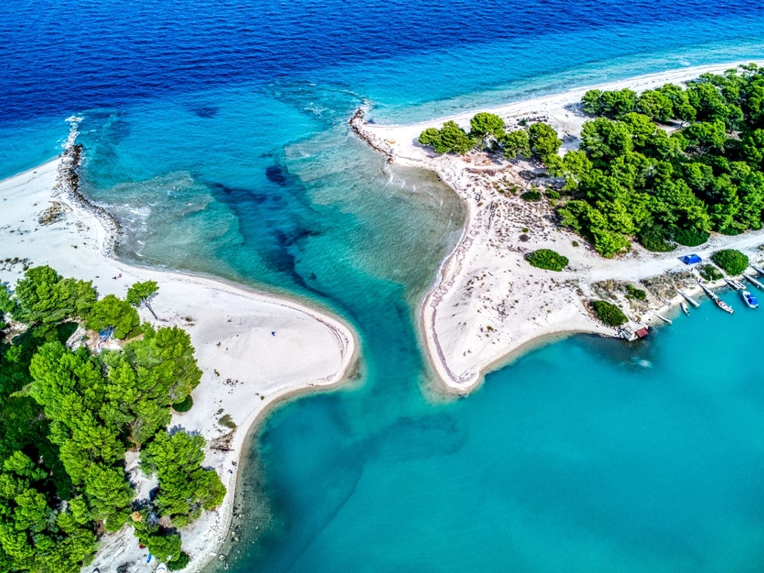 Luftfoto af en smal turkis kanal, der skærer gennem en hvid sandtange, flankeret af grønne træer og en bådebro.