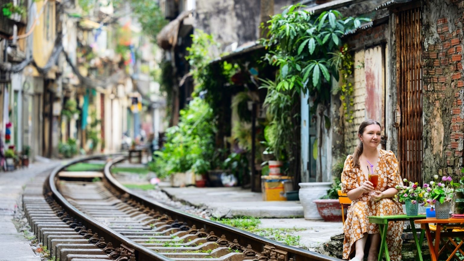 En kvinde sidder ved et bord ved siden af jernbanespor, der buer gennem en smal bygade omgivet af gamle bygninger. Hanoi, Vietnam
