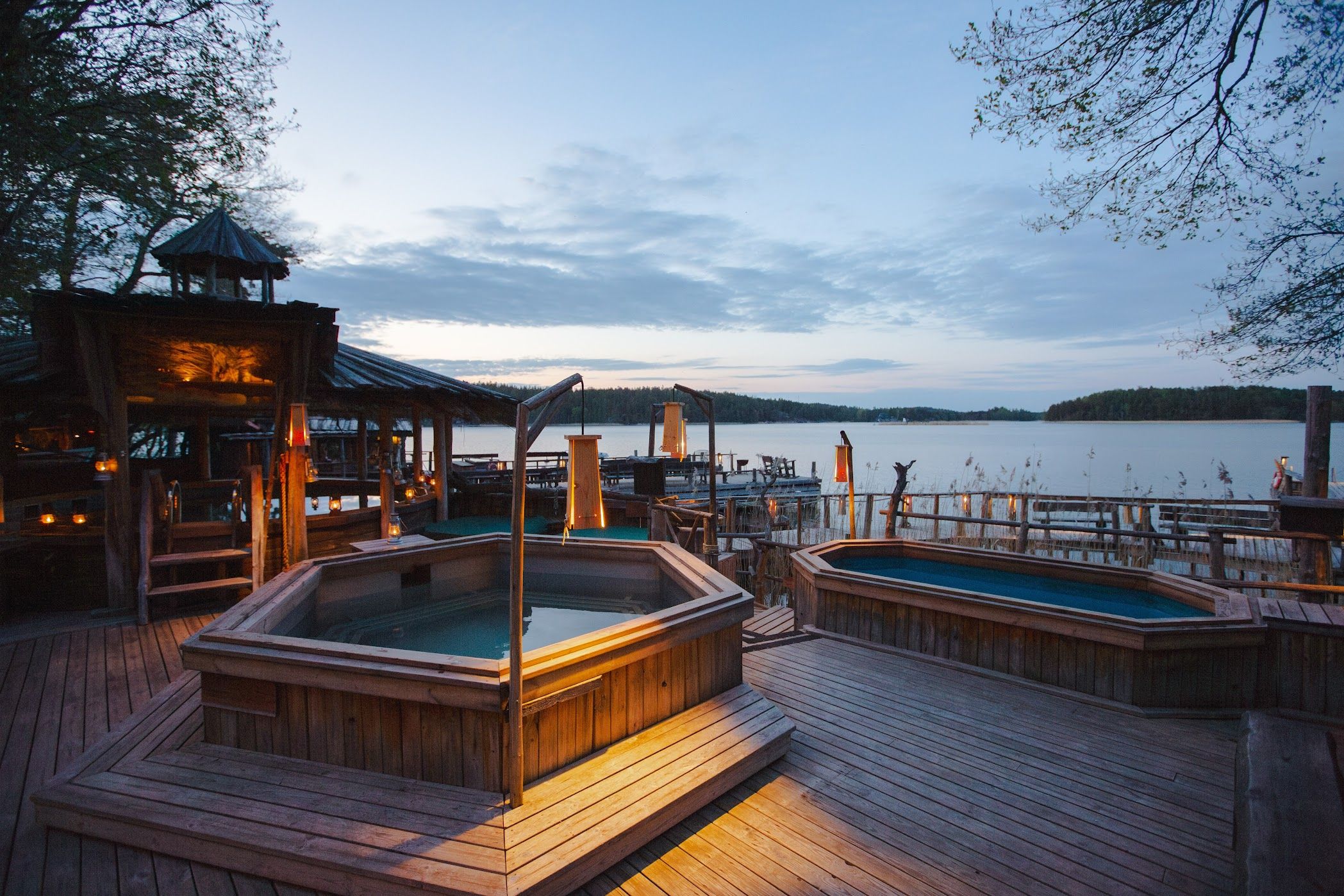 Terrasse ved søen med to træbade og en oplyst pavillon i skumringen.