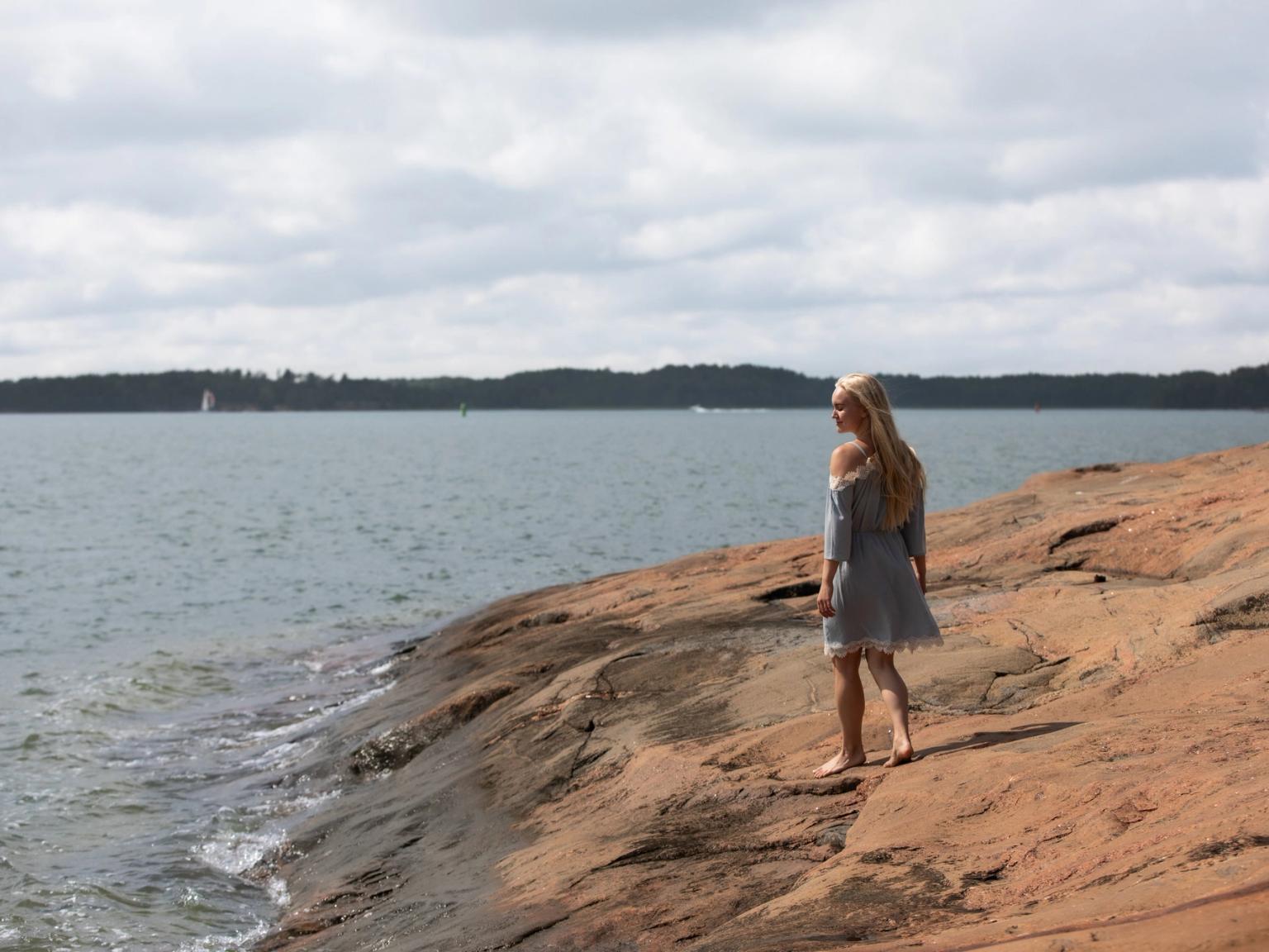 Vaaleahiuksinen nainen kevyessä mekossa kävelee paljain jaloin punaisilla kallioilla veden äärellä ja katsoo olkansa yli taaksepäin.
