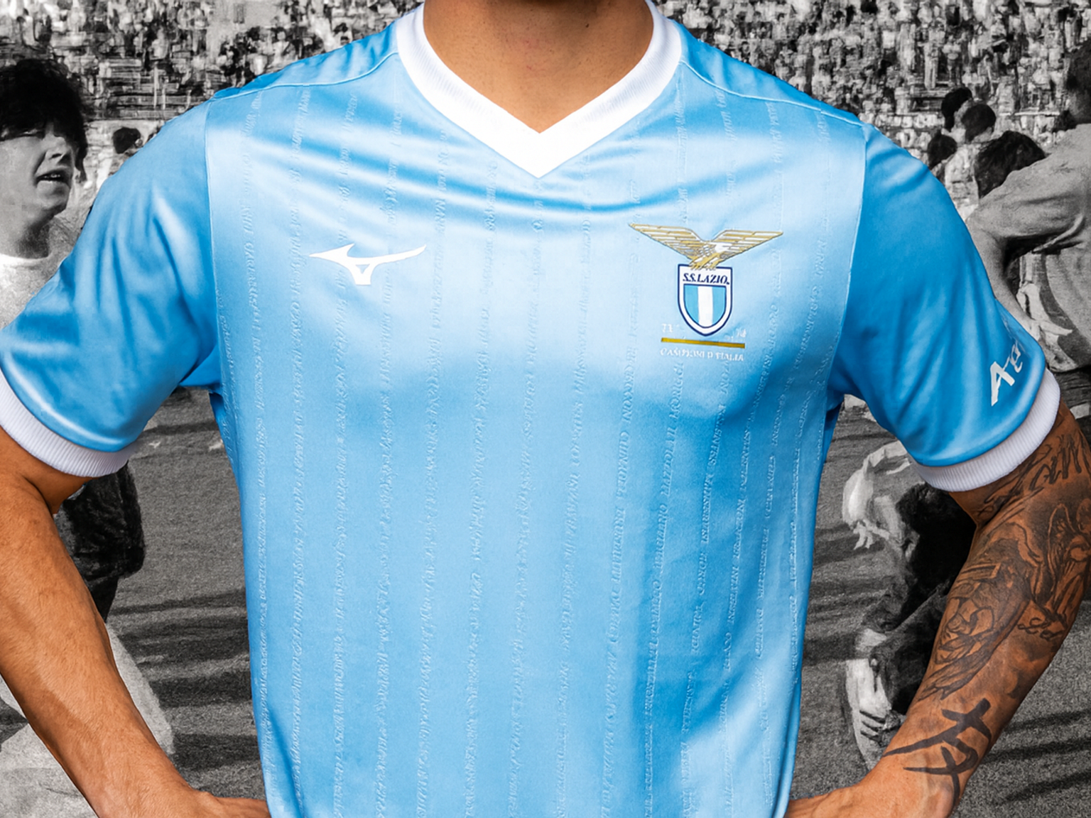 Närbild av en ljusblå Lazio fotbollströja med klubbens emblem och Mizuno-logotyp, buren av en person med tatuerad arm, överlagrad på en bleknad svartvit folkmassa.