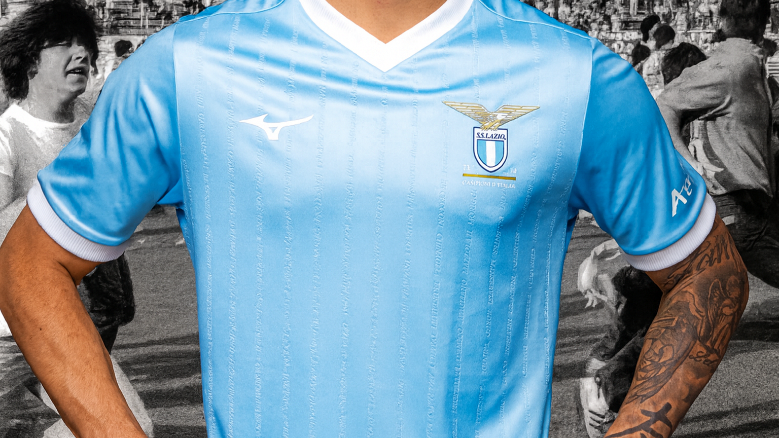 Close-up of a light blue Lazio soccer jersey with club crest and Mizuno logo, worn by a person with a tattooed arm, overlaid on a faded black and white crowd.