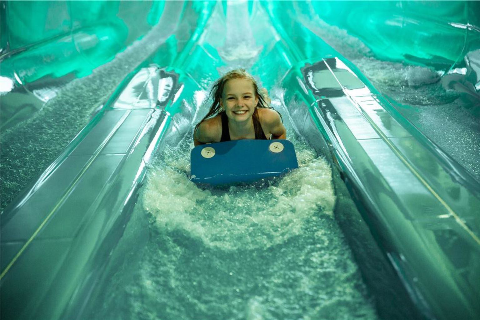 En smilende jente på et blått bodyboard sklir ned en vannpark sklie med sprutende vann. Lalandia Resort Søndervig