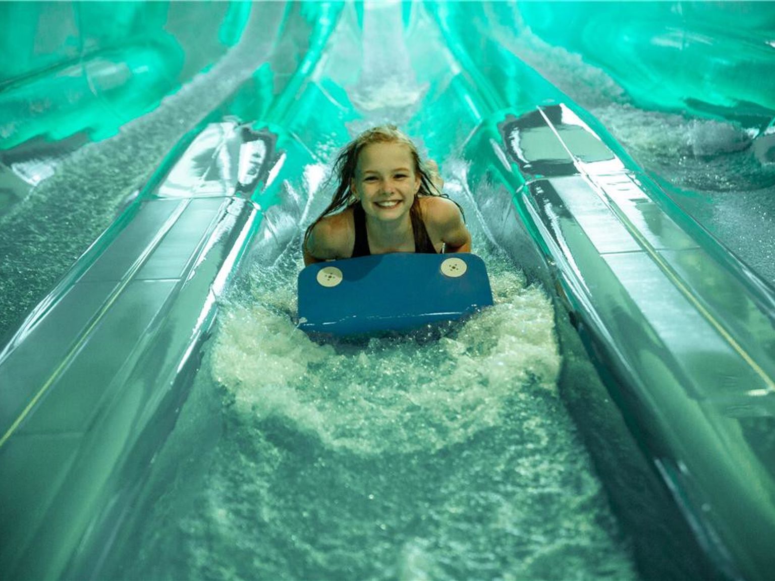 En smilende pige på et blåt bodyboard glider ned ad en vandpark rutsjebane med sprøjtende vand. Lalandia Resort Søndervig
