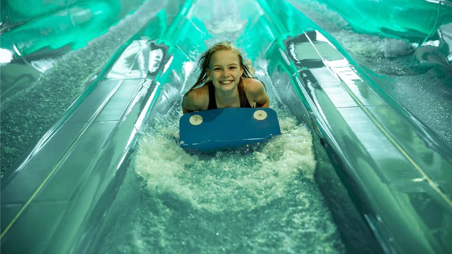 En smilende jente på et blått bodyboard sklir ned en vannpark sklie med sprutende vann. Lalandia Resort Søndervig