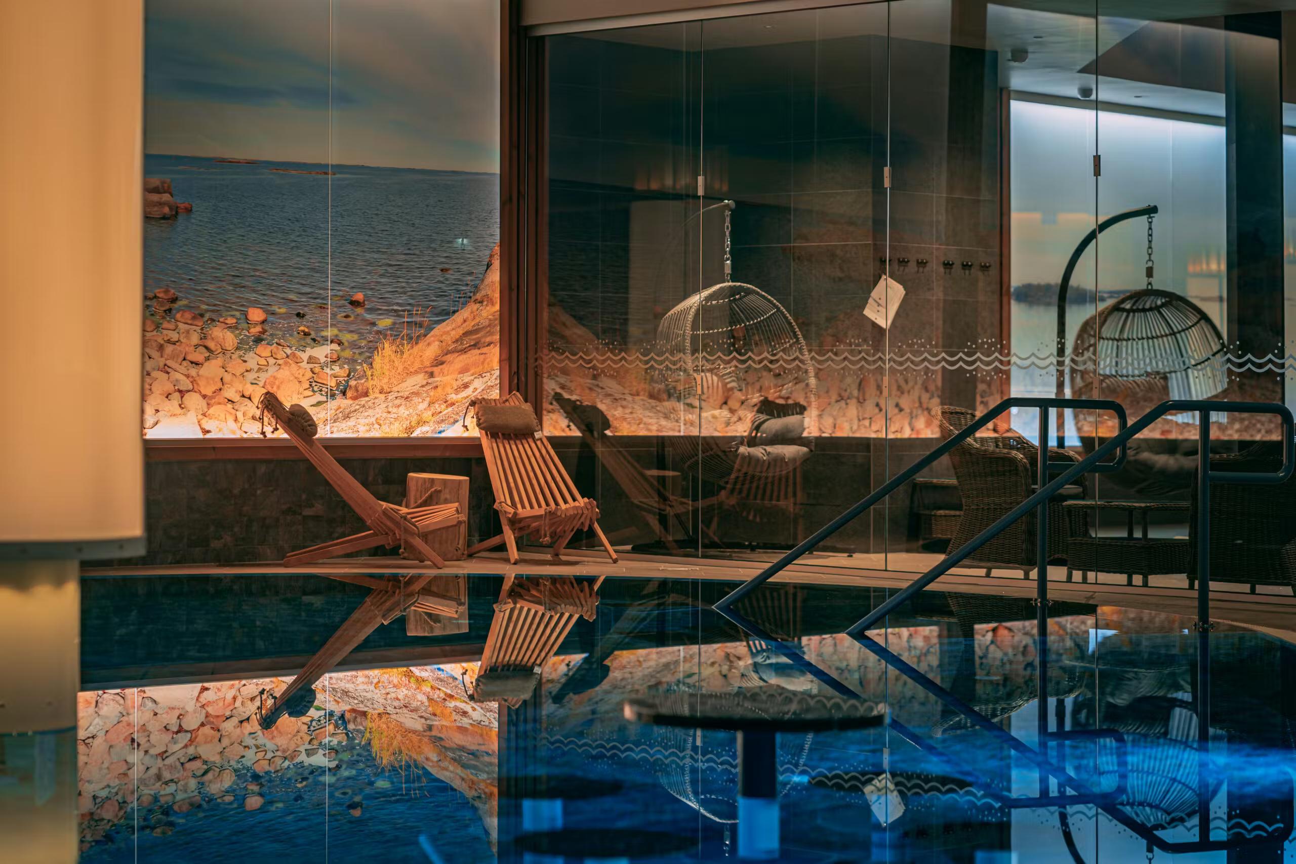 Indoor pool with a scenic coastal mural, a wooden lounge chair, and an anchor by the water's edge.